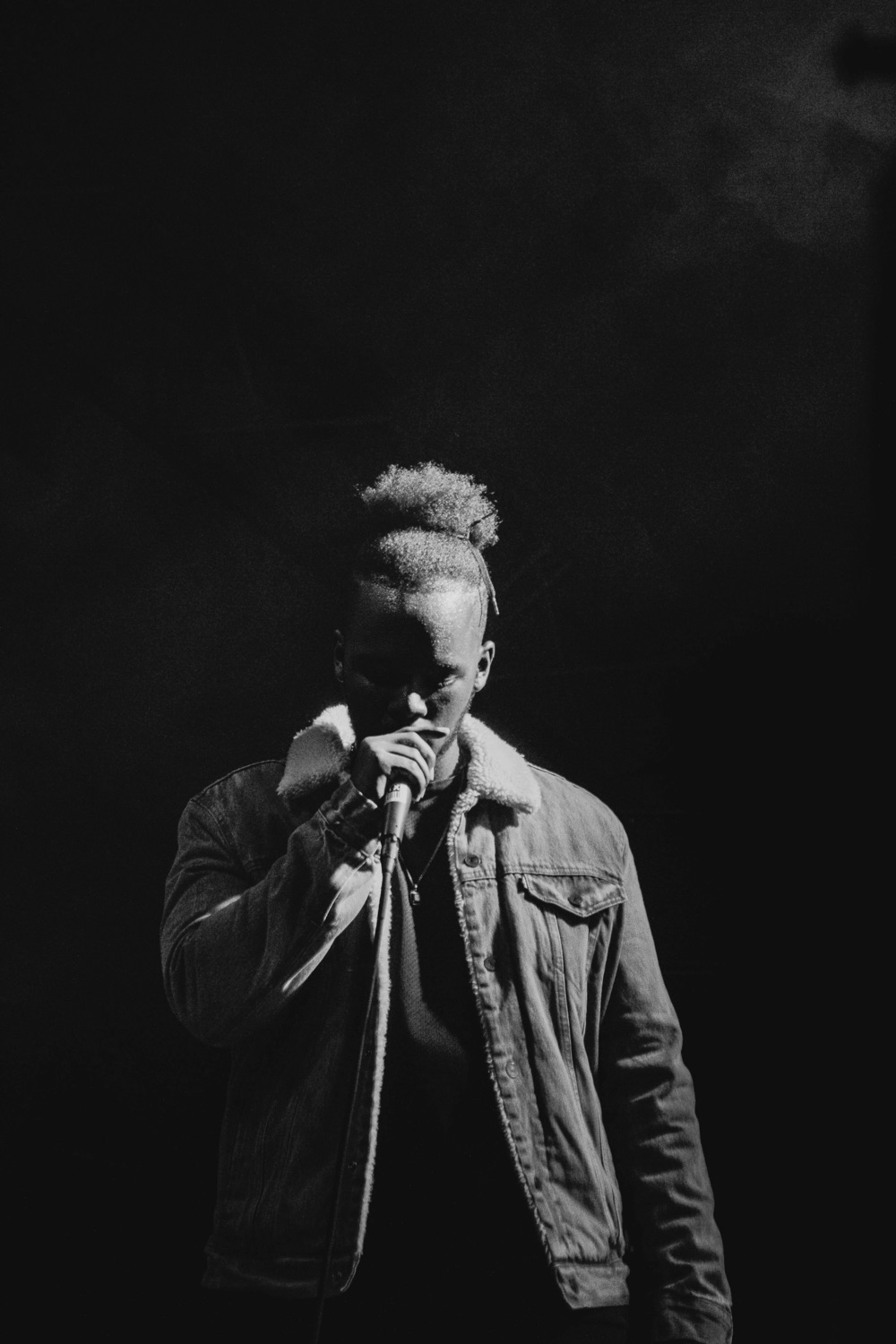 Rapper in a denim jacket singing into a microphone under a spotlight, in black and white